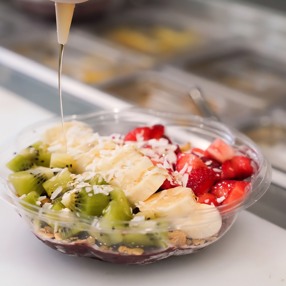 Fruit bowl topped with kiwi, strawberries, banana, shredded coconut, and syrup being poured over it.