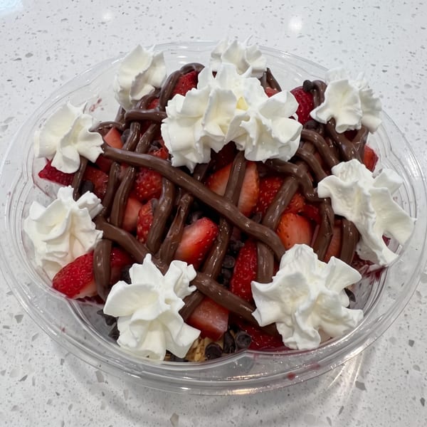 A clear plastic bowl filled with sliced strawberries, chocolate drizzle, and whipped cream.