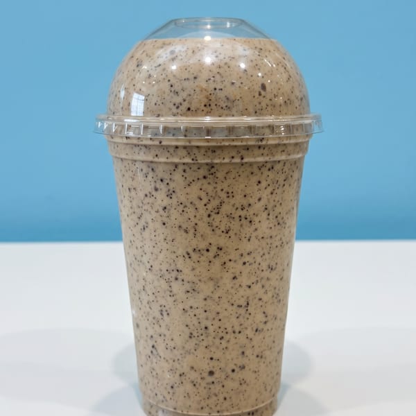 Creamy, chocolate-speckled milkshake in a clear domed plastic cup on a white surface against a blue background.