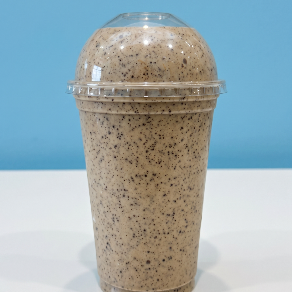 Creamy, chocolate-speckled milkshake in a clear domed plastic cup on a white surface against a blue background.