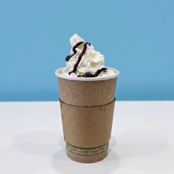 Soft-serve ice cream topped with whipped cream and chocolate sauce in a recyclable paper cup against a blue background.