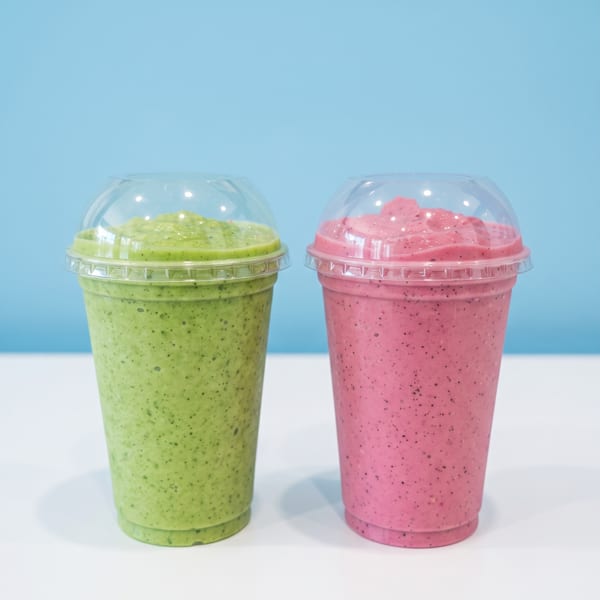 Two colorful smoothies, green and pink, in clear-lidded plastic cups against a bright blue background.